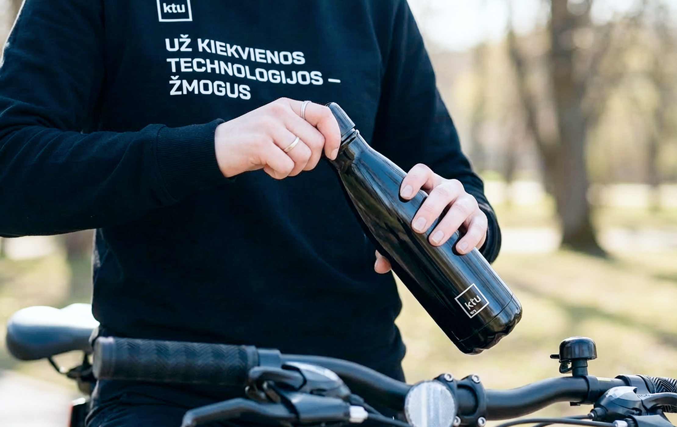 Žmogus laiko daugkartinio naudojimo gertuvę su KTU logotipu &scaron;alia dviračio. Ant megztinio matomas KTU &scaron;ūkis &bdquo;Už kiekvienos technologijos &ndash; žmogus&ldquo;. Vizualas perteikia tvarumo principus KTU konferencijose, skatinant naudoti daugkartinius daiktus ir rinktis aplinkai draugi&scaron;ką judumą.