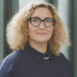 Prof. dr. Violeta Motūzienė, Vilniaus Gedimino technikos universiteto pastatų energetikos katedros vedėja, konferencijos prane&scaron;ėja. Portretinė nuotrauka &ndash; moteris su &scaron;viesiais garbanotais plaukais ir akiniais, dėvinti tamsią palaidinę. Ji žiūri į kamerą. Fonas &ndash; &scaron;viesus.